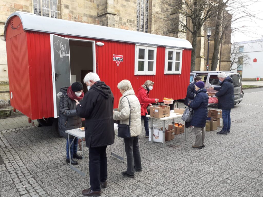 Vor dem roten CVJM-Bauwagen sind zwei Tische aufgebaut. Zwei CVJM-Mitglieder verkaufen dort Orangen an vier Kunden.